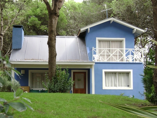 A two-story blue chalet is surrounded by greenery and tall trees. A metal roof and a balcony with white railings highlight the architecture. Large windows allow natural light to fill the interior, while a welcoming entrance features a wooden door.