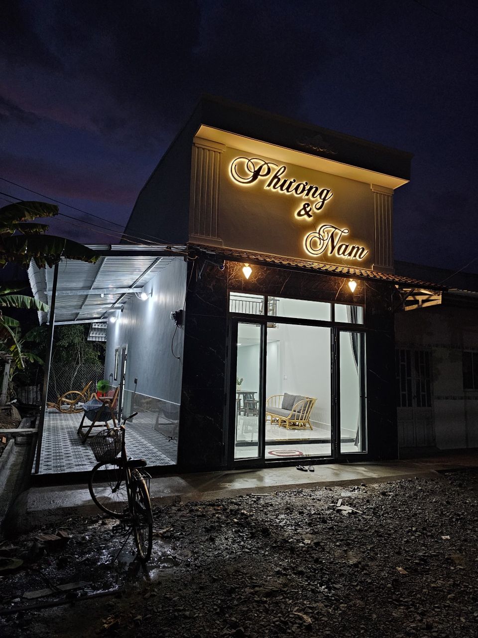 The exterior of a stylish building is illuminated at night, showcasing the name 'Phuong & Nam' in soft lighting. Large glass doors create a welcoming entrance, while outdoor seating is visible under a covered area, surrounded by a gravel path and greenery.