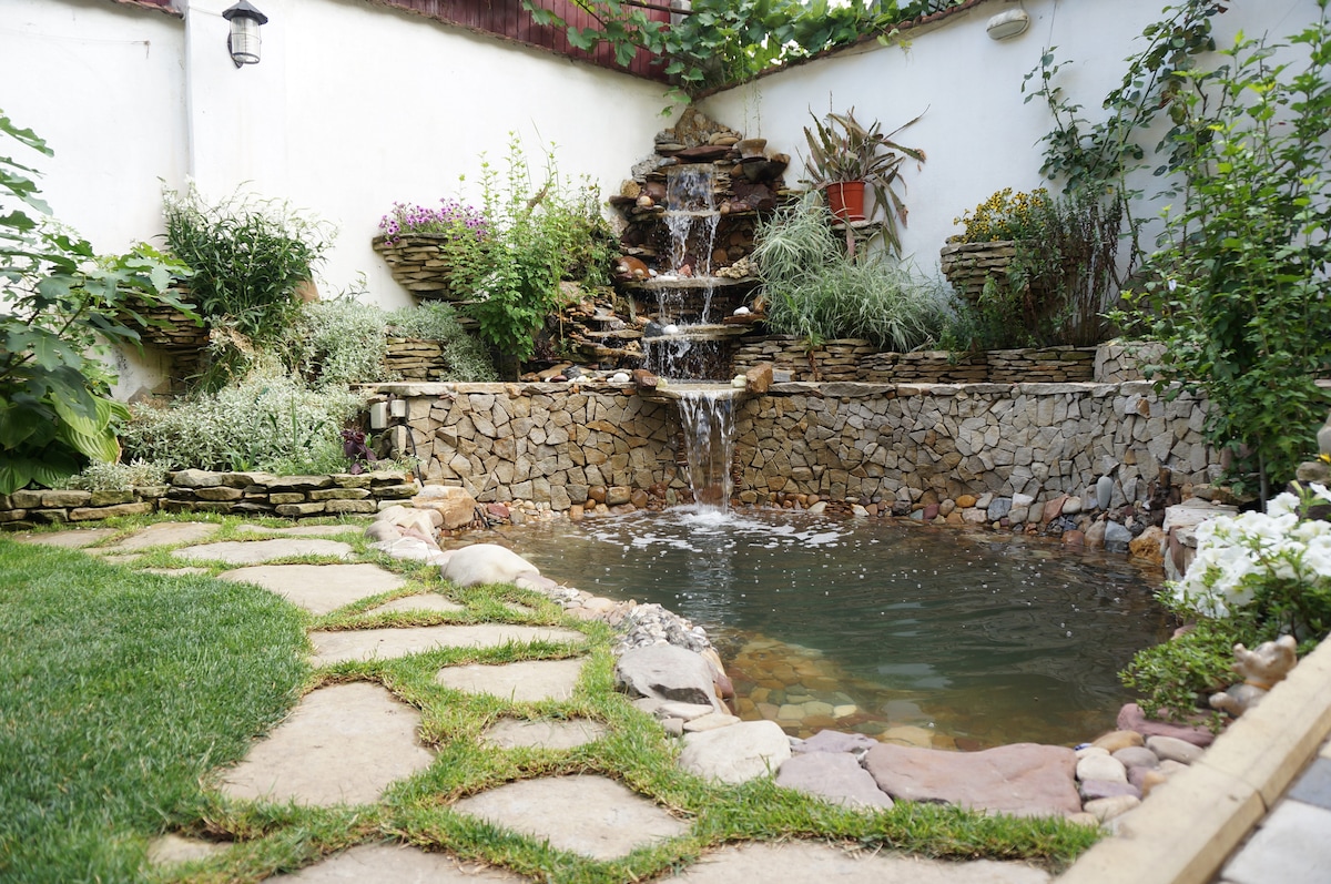 A serene garden area is highlighted by a natural stone pond surrounded by lush greenery and vibrant flowers. A gently flowing water feature adds a calming sound, while the stone pathway leads through the well-maintained landscape.