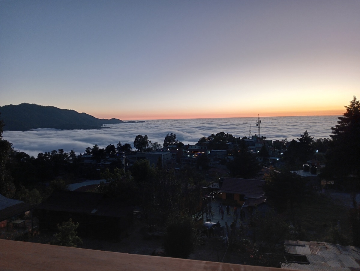 A serene view of a cloud-covered valley at sunset, where the horizon is painted with warm hues. The silhouette of distant mountains frames the scene, while a peaceful village is nestled among trees below, offering a glimpse of daily life.