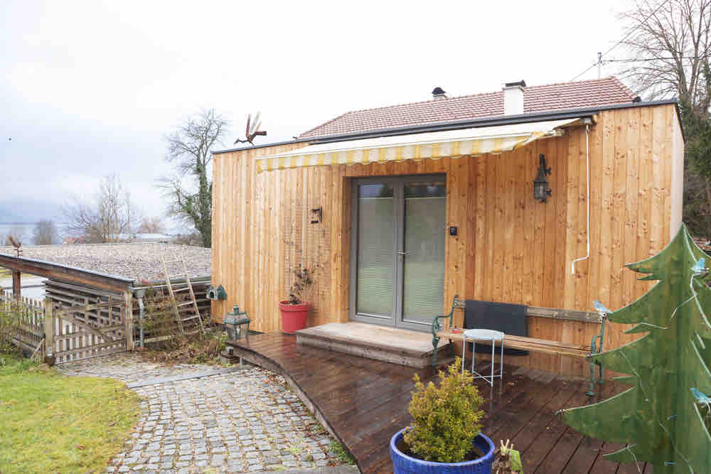 A charming tiny house constructed with wooden paneling is shown, featuring a covered entrance and a small patio. A garden area with potted plants and a decorative tree is visible, along with a sloped pathway leading to the entrance.