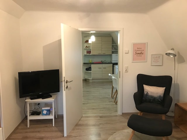 A welcoming living area is shown, featuring a television and an armchair with wooden legs. A view through an open door reveals a modern kitchen with light cabinetry. Natural light enters the space, creating a bright and inviting atmosphere.