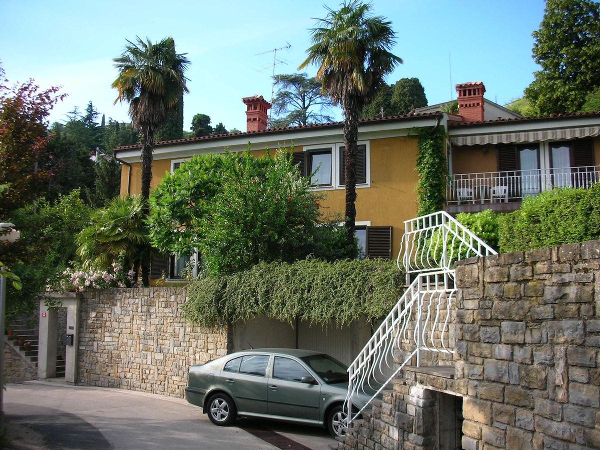 A family house is set within a peaceful garden, featuring a stone wall and palm trees. A car is parked in the driveway, and a stairway leads to a balcony adorned with greenery. The exterior showcases earthy tones and tiled roofing.