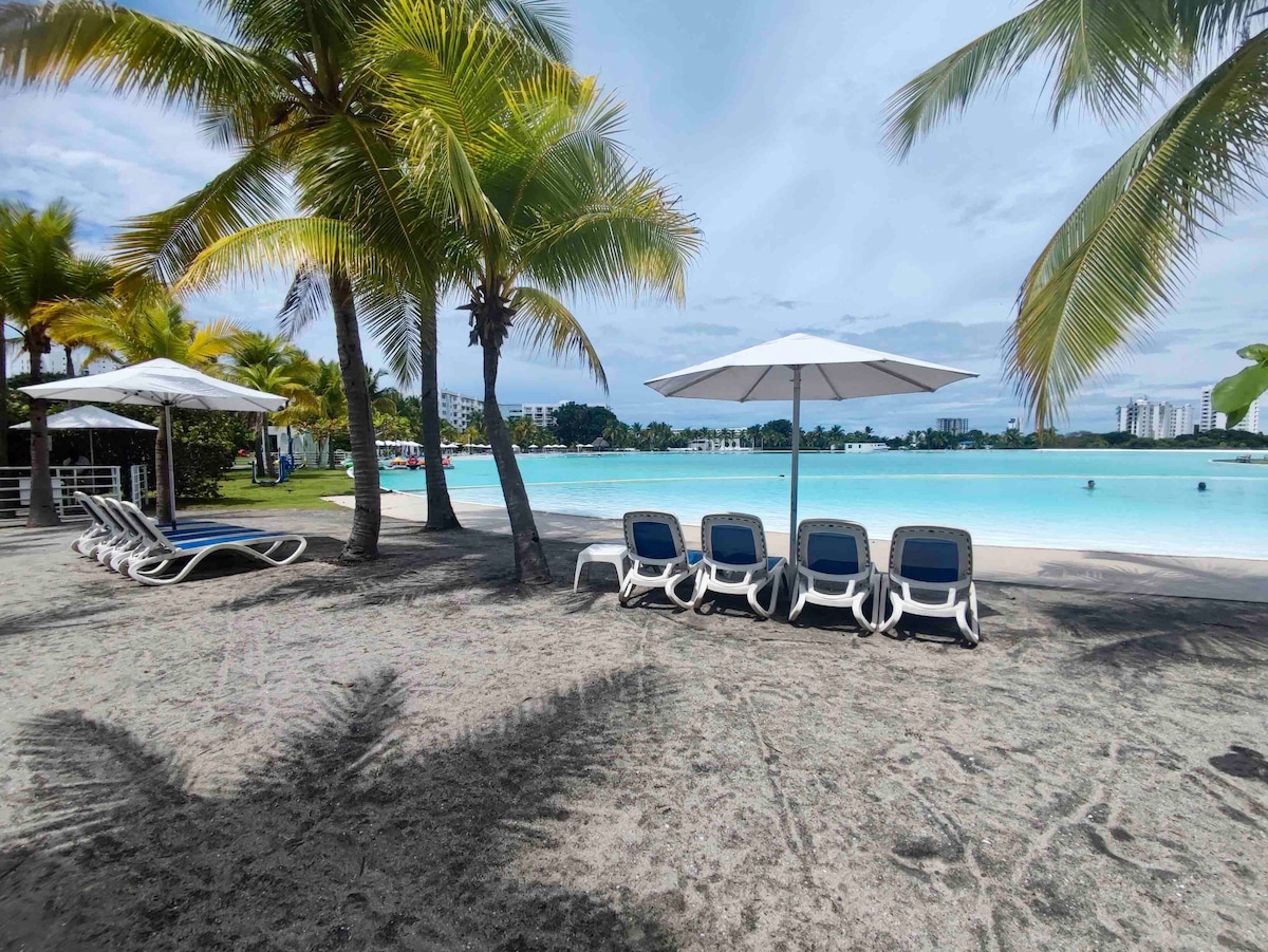 A sandy shoreline is framed by tall palm trees, providing shade with white umbrellas. Several lounge chairs are arranged in a row, facing a large artificial lagoon. The clear water and gentle waves create a serene atmosphere, inviting relaxation by the water's edge.