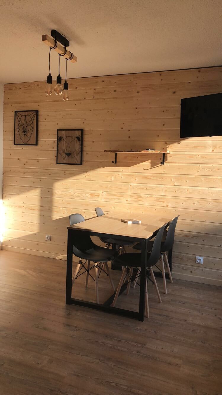 A dining area is highlighted with a modern table surrounded by four chairs. The wooden wall adds warmth, complemented by decorative wall art. Natural light filters in, creating a serene environment. A mounted TV is positioned above a sleek shelf.