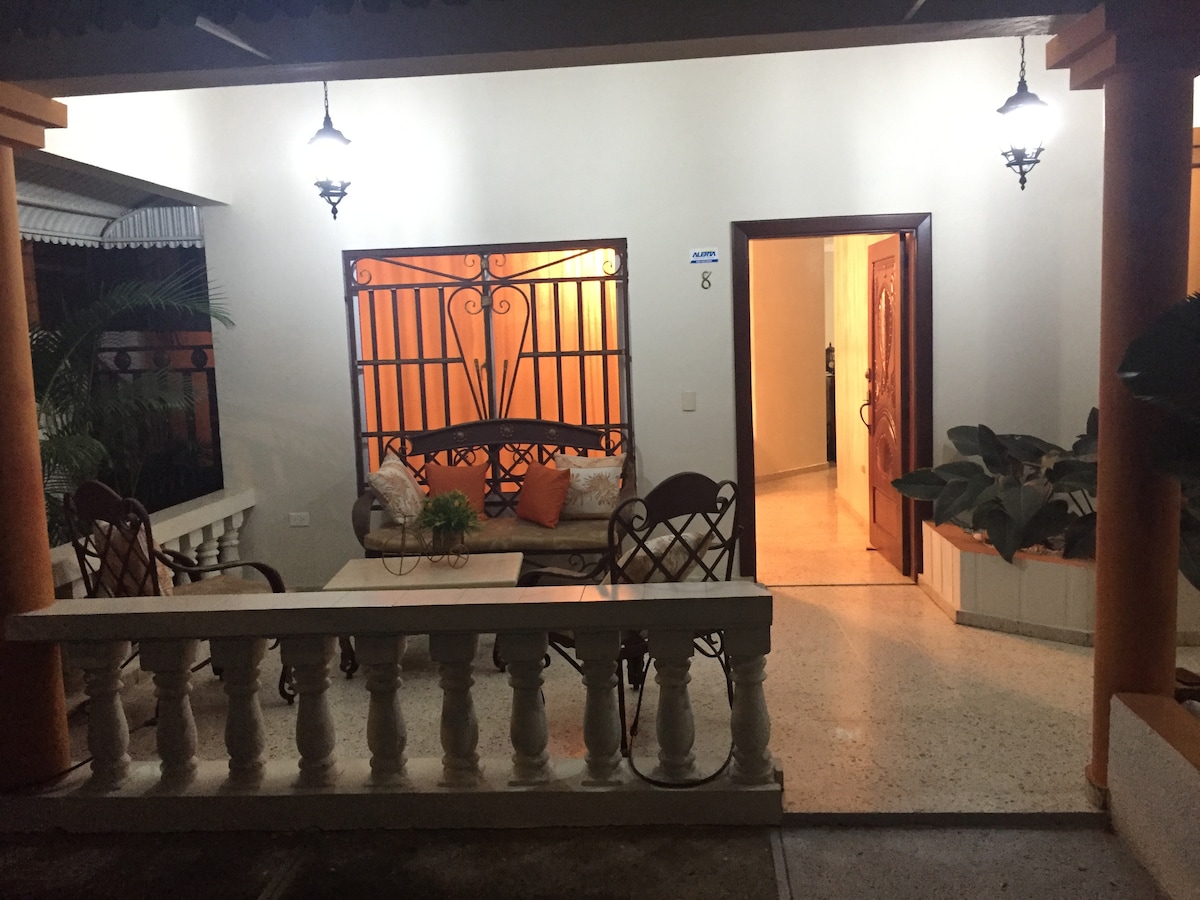 An inviting porch area is visible, featuring a comfortable seating arrangement with a small table. Decorative plants are placed nearby, and traditional lanterns illuminate the space, creating a warm atmosphere. The entrance to the interior is framed by a sturdy door and visible through an open archway.