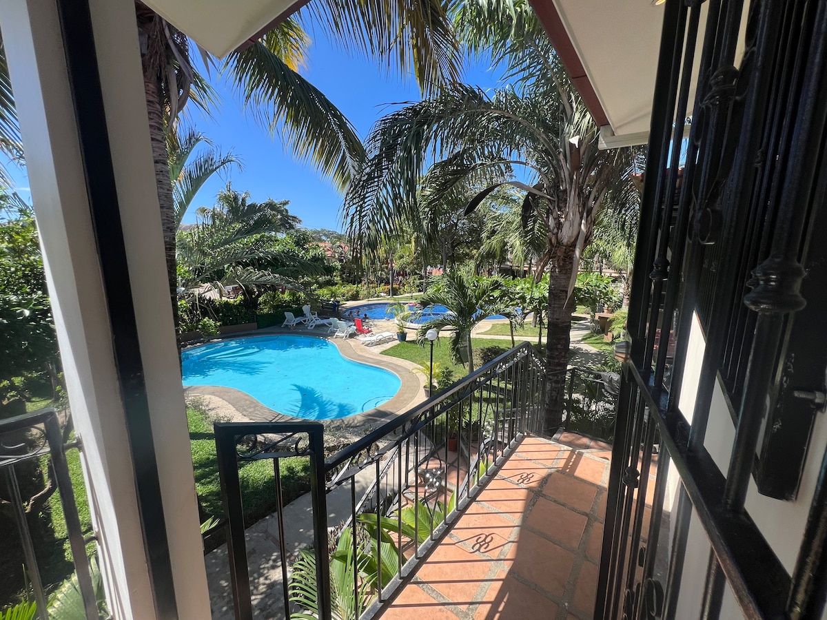 A view from a balcony features a vibrant garden with palm trees and lush vegetation surrounding a clear swimming pool. Lounge chairs are positioned near the water's edge, offering a serene outdoor space. Bright blue skies create a tranquil atmosphere.