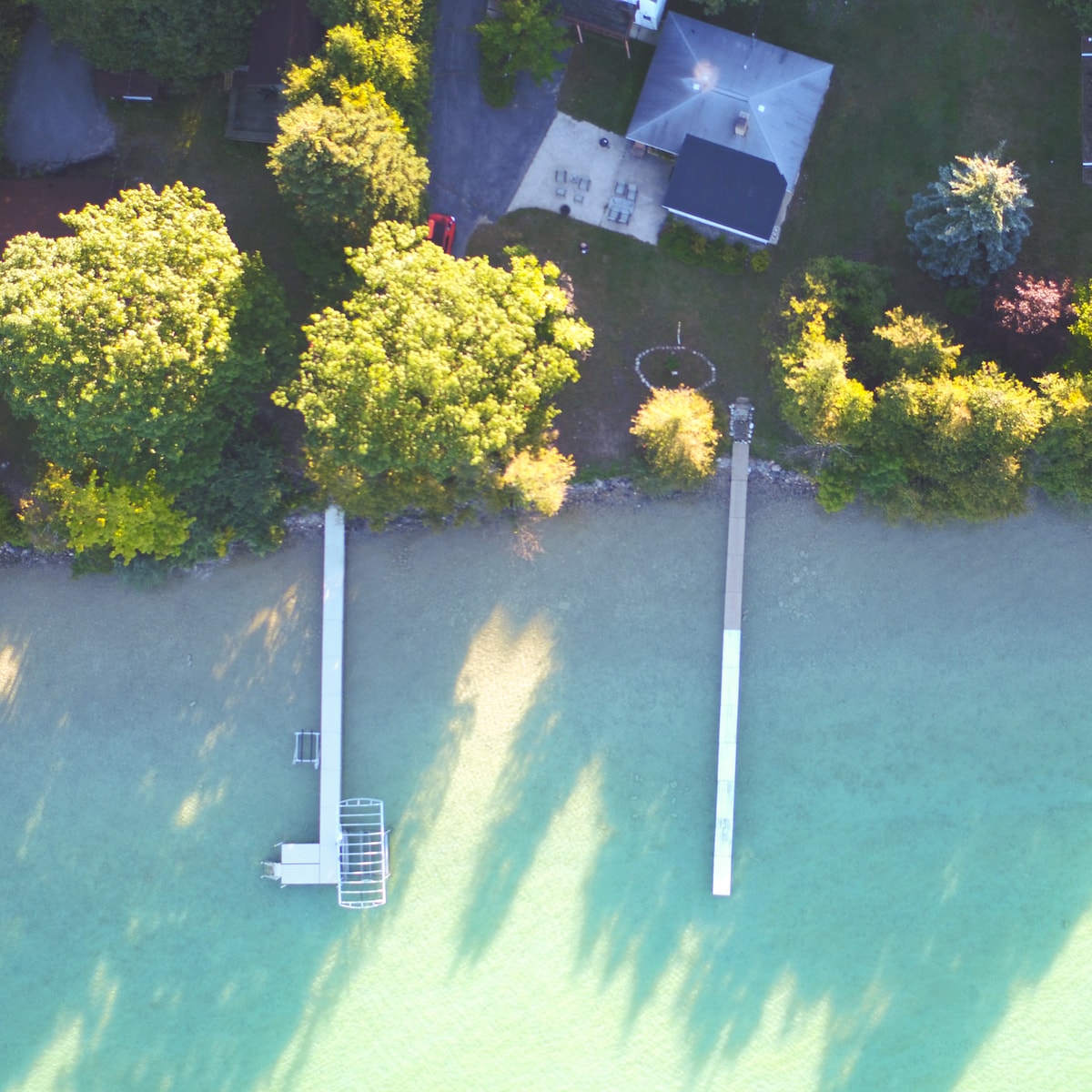 An aerial view captures the serene Torch Lake shoreline, featuring two docks extending into the clear water. Lush green trees surround the area, and a grassy lawn can be seen adjacent to the cottage, providing plenty of outdoor space for relaxation.