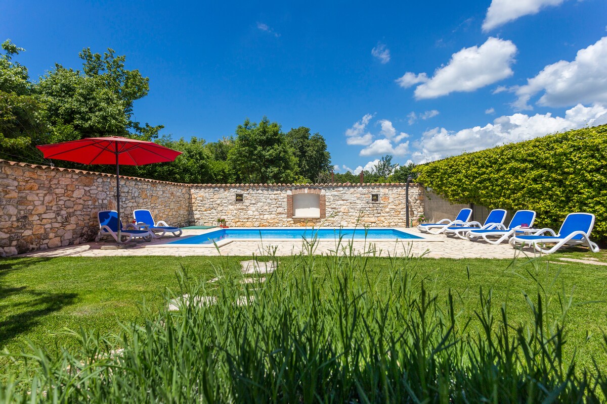 An outdoor pool area is surrounded by lush greenery and a stone wall, providing privacy. Blue lounge chairs line one side, with a red umbrella offering shade. The sky is clear with a few scattered clouds, creating a bright and open atmosphere.