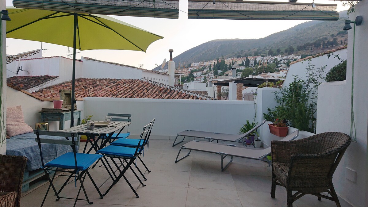 A spacious terrace is equipped with a dining table and blue chairs, complemented by a shaded area with a large umbrella. Lounge chairs are positioned for relaxation, while potted plants add greenery. Views of the rooftops and surrounding mountains are visible in the background.