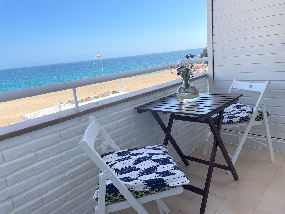 A balcony area features a small wooden table with two white folding chairs, each adorned with patterned cushions. A vase with fresh flowers is placed on the table. The backdrop showcases a panoramic view of the clear blue sea and sandy beach.