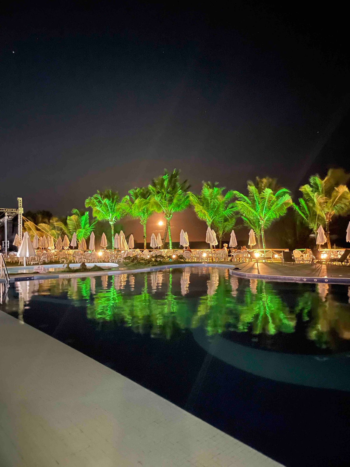 The nighttime scene features a shimmering pool illuminated by green lights, surrounded by palm trees. Umbrellas are positioned around the pool area, reflecting softly in the water. A warm glow from the nearby lighting enhances the inviting atmosphere of the space.