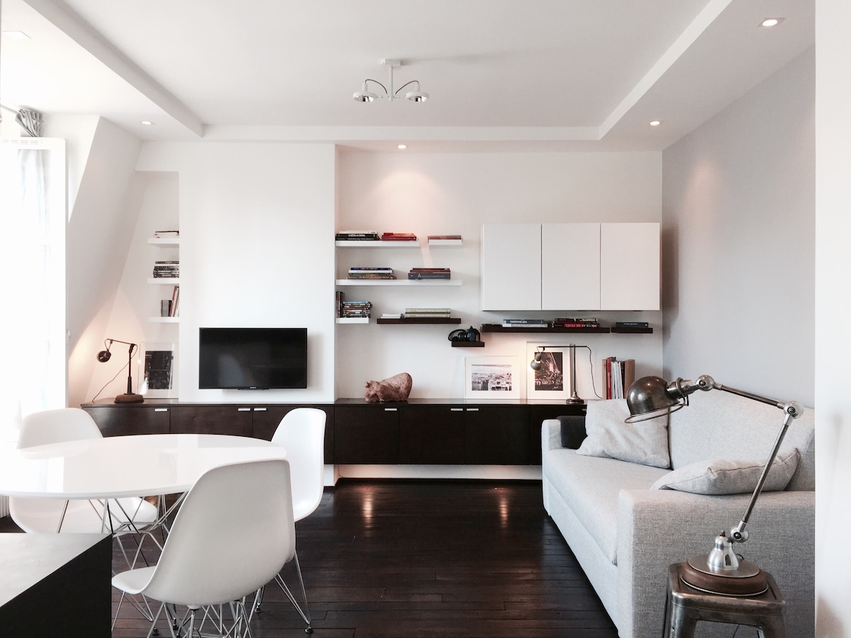 The living area features modern shelving with books and decorative items, complemented by a sleek flat-screen TV. A cozy gray sofa offers seating, while a round dining table with white chairs occupies the space, illuminated by soft overhead lights.