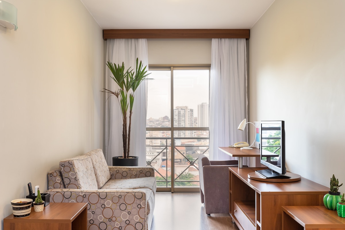 A bright living area is highlighted by large windows that offer city views. A textured sofa and desk, accompanied by a chair, provide comfortable seating. Decorative plants and modern furnishings add to the space's appealing decor.