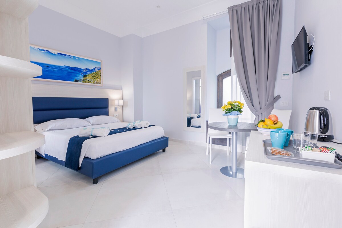 A spacious room features a large bed with soft linens, complemented by a minimalist design and light blue walls. A small table is set with a fruit bowl and beverages. Natural light fills the space, and a scenic wall art piece adds a touch of elegance.
