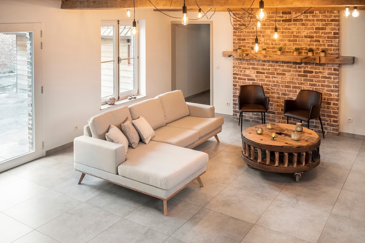 A spacious living area is presented, featuring a light-colored sectional sofa with decorative cushions. A wooden coffee table made from an old mill wheel is centered in the room. Exposed brick walls and industrial light fixtures enhance the ambiance. Large windows provide natural light.
