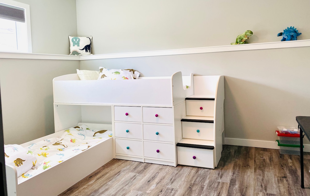 A kid-friendly bedroom is equipped with a white bunk bed, featuring a lower bed with a fun dinosaur-themed bedding. Colorful storage drawers are present beneath the upper bed, and a small staircase with playful knobs provides access to the upper sleeping area.