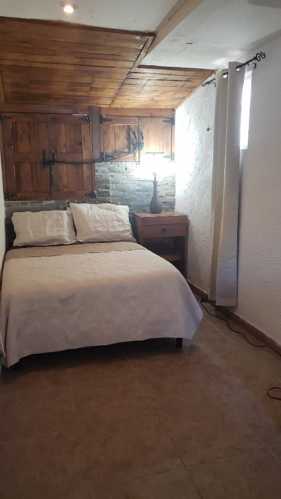 A cozy sleeping area features a simple bed with light bedding and two pillows. A bedside table is positioned next to the bed, accompanied by a lamp. Wooden accents and stone walls contribute to the room's charming character, with natural light entering through a small window.