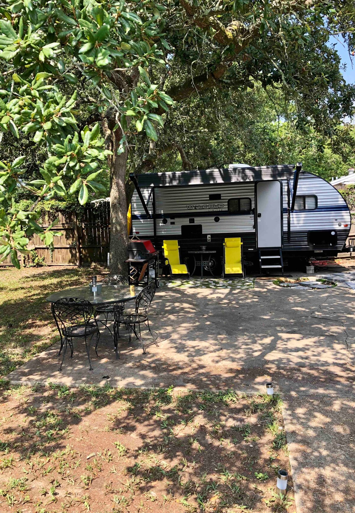 A compact campsite is showcased, featuring a sleek camper set against a backdrop of trees. A black outdoor table with four chairs is positioned on a concrete patio, surrounded by grass and shaded by the branches of a nearby tree.