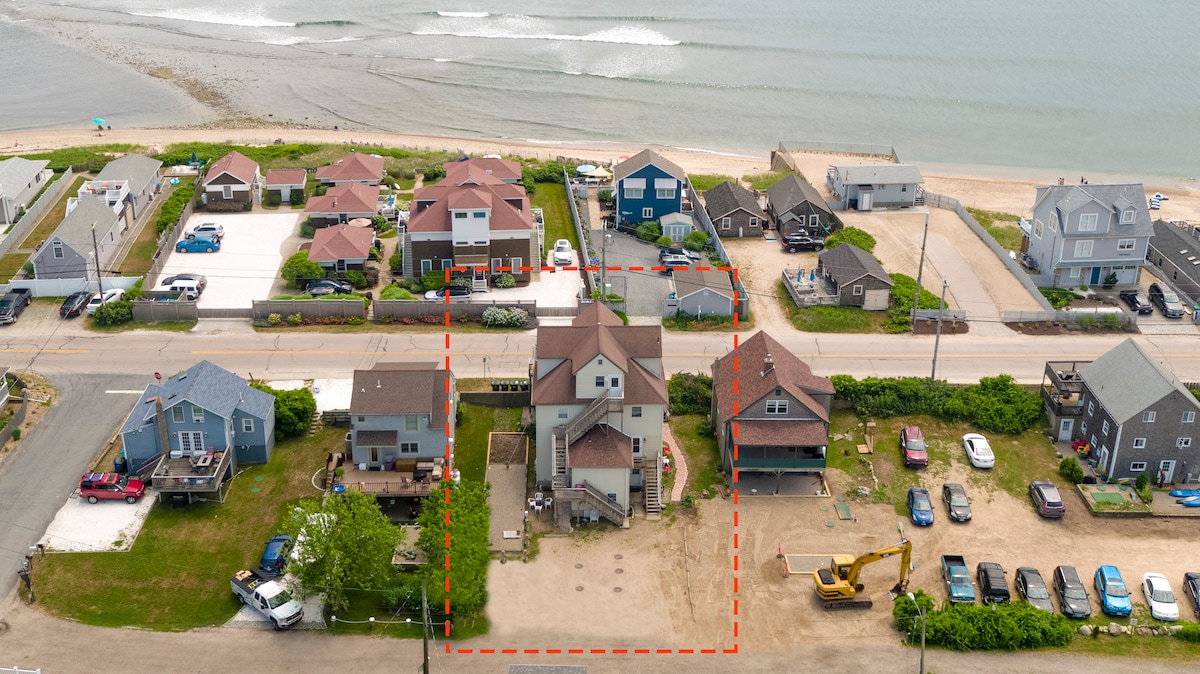 An aerial view captures a coastal area featuring a collection of homes, parking spaces, and beach access. The layout showcases multiple residential buildings arranged along a road, with the ocean visible in the background. Greenery surrounds the properties, adding to the tranquil setting.