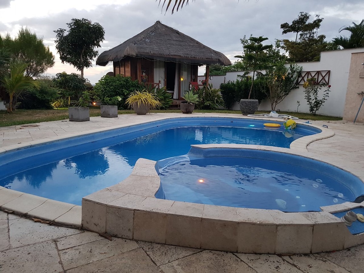 A clear swimming pool is positioned in the foreground, featuring a jacuzzi section. Surrounding the pool are well-maintained gardens with tropical plants. In the background, a charming bungalow with a thatched roof can be seen, adding to the serene outdoor space.