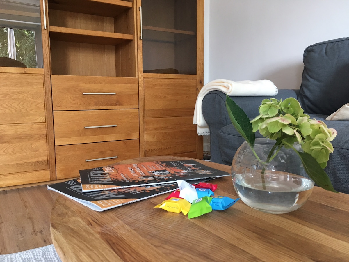 A wooden coffee table displays a small glass vase with green foliage and colorful candy wrappers alongside magazines. A gray sofa and wooden shelving unit are visible in the background, contributing to a cozy, organized living space.