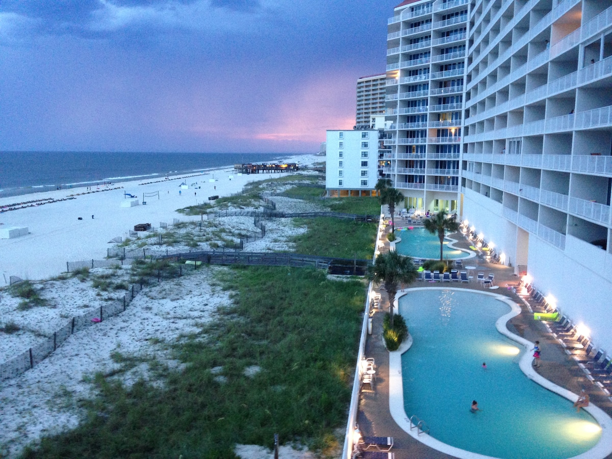 The view displays the serene beachfront along the Gulf of Mexico, showcasing soft waves and a sandy shore. A resort-style pool is visible, surrounded by lounge chairs and palm trees, while the evening sky is painted with gentle hues of purple and blue. Nearby buildings line the coast.