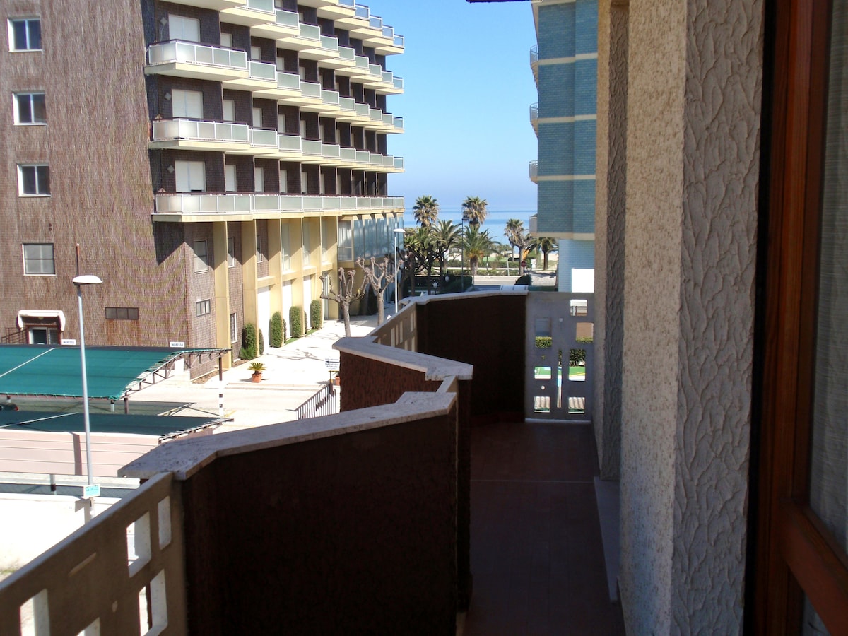 A balcony is visible, offering a view of nearby buildings and palm trees. The bright blue sky and distant sea are seen in the background. The balcony features a textured railing and tiled flooring, creating an open and airy space.