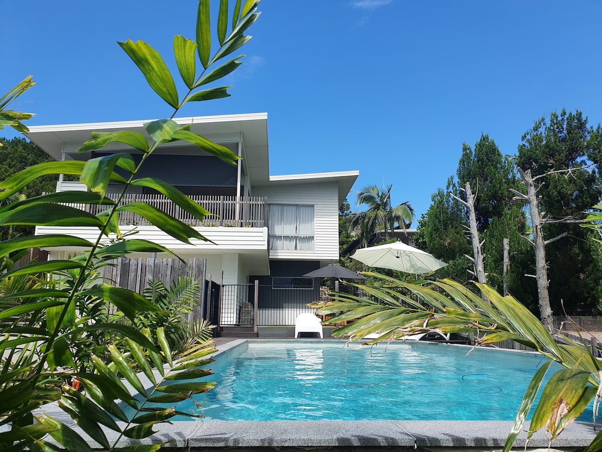 A modern two-story home is visible, set against a clear blue sky. A swimming pool is positioned in the foreground, surrounded by tropical greenery, with a sun lounger and umbrella nearby. The balcony offers a view of the lush surroundings.