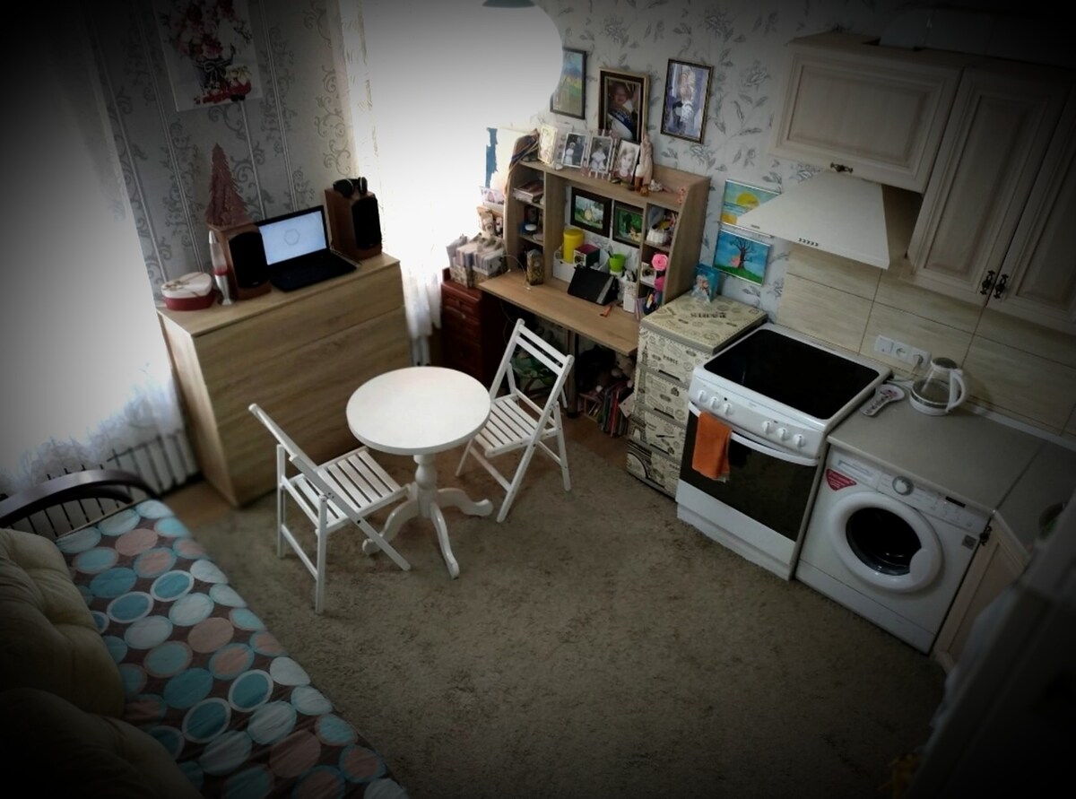 A compact living area is showcased, featuring a small round table with two white chairs. A light-colored sideboard stands against the wall, decorated with various items. A washing machine is visible next to the kitchen area, with soft beige carpeting covering the floor.