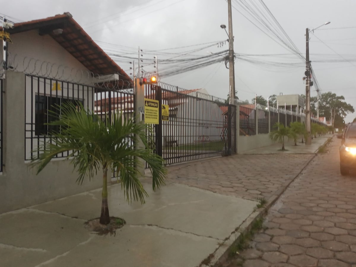 The exterior of the property is shown, featuring a gated entrance with security features. A paved pathway leads alongside a row of small palm trees. Electrical wiring is visible overhead, and a vehicle is parked near the entrance, indicating accessibility to the location.