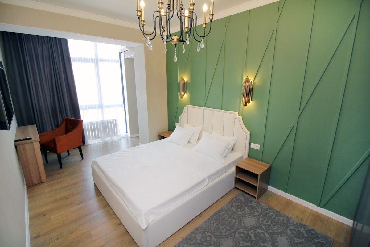 A modern bedroom is presented with a spacious bed positioned against a green accent wall. Soft lighting from a chandelier adds warmth, while a nearby window is partially covered by dark curtains. A simple desk and an orange armchair complete the inviting space.