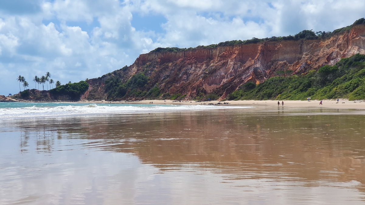 A sandy beach stretches alongside a vibrant coastline, with rugged cliffs rising in the background. Lush greenery is present atop the cliffs, and gentle waves lap at the shore, reflecting the clouds in the sky. People are seen walking along the water's edge.