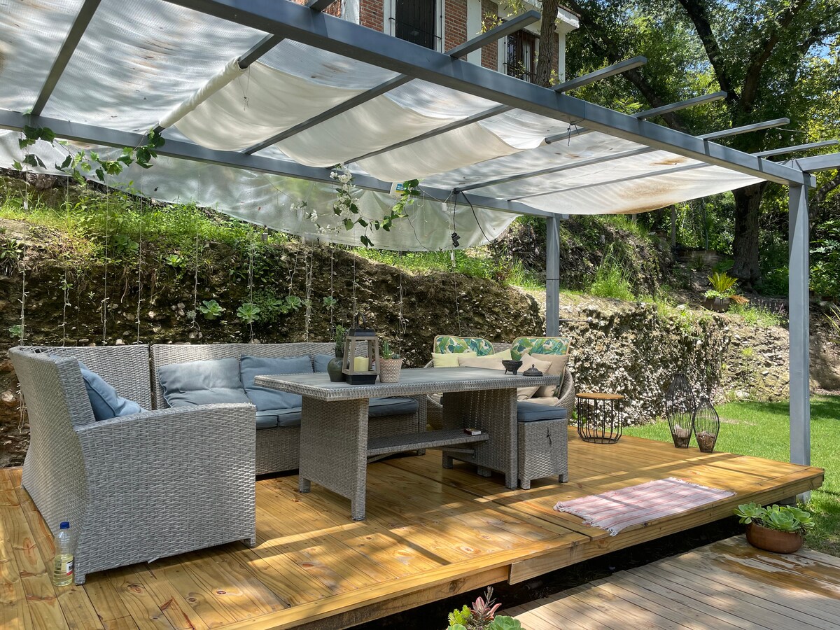 A shaded outdoor seating area is presented with a wooden deck, featuring a comfortable table and chairs. Greenery hangs above, creating a tranquil space. A soft rug lies partially on the floor, enhancing the inviting ambiance of the setting.