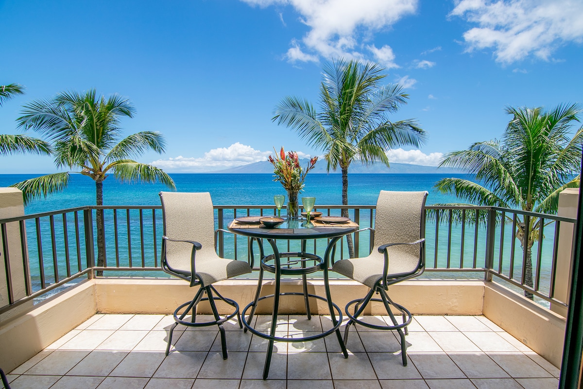 A private lanai is depicted, featuring a round glass-top table and two high-backed chairs. Fresh flowers are arranged in the center, with vibrant blue ocean views and swaying palm trees visible in the background under clear skies.