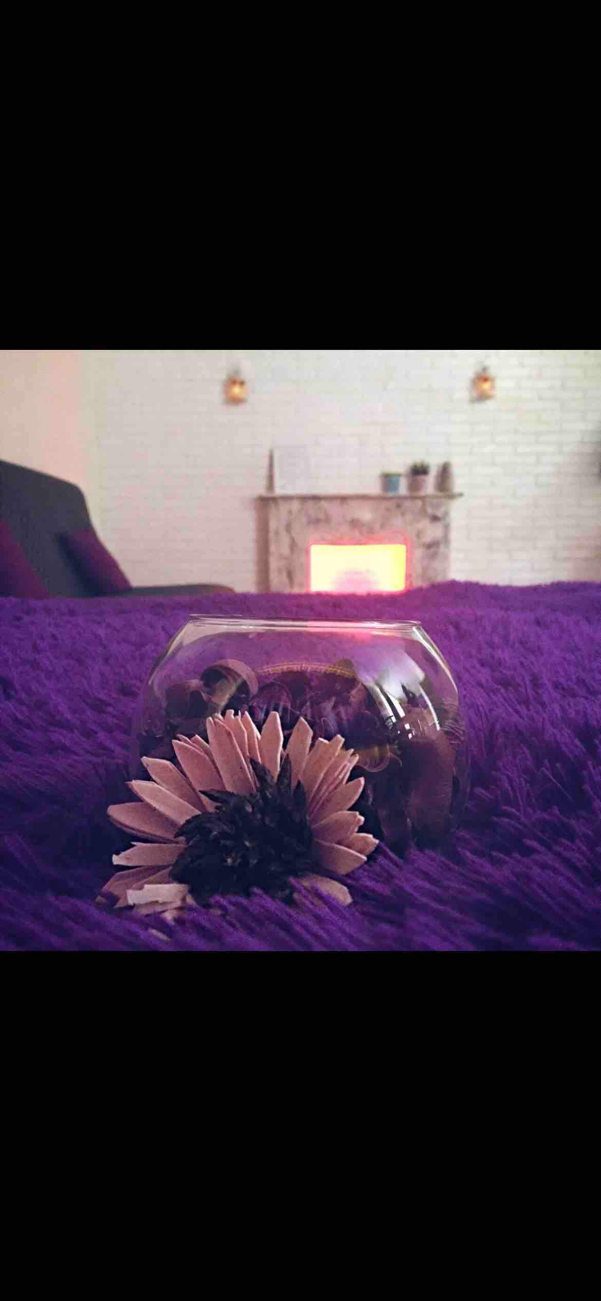 A close-up view of a glass vase containing dried flowers is positioned on a vibrant purple surface. In the background, a soft glow from a fireplace is visible, adding warmth to the setting. The overall composition suggests a serene atmosphere.