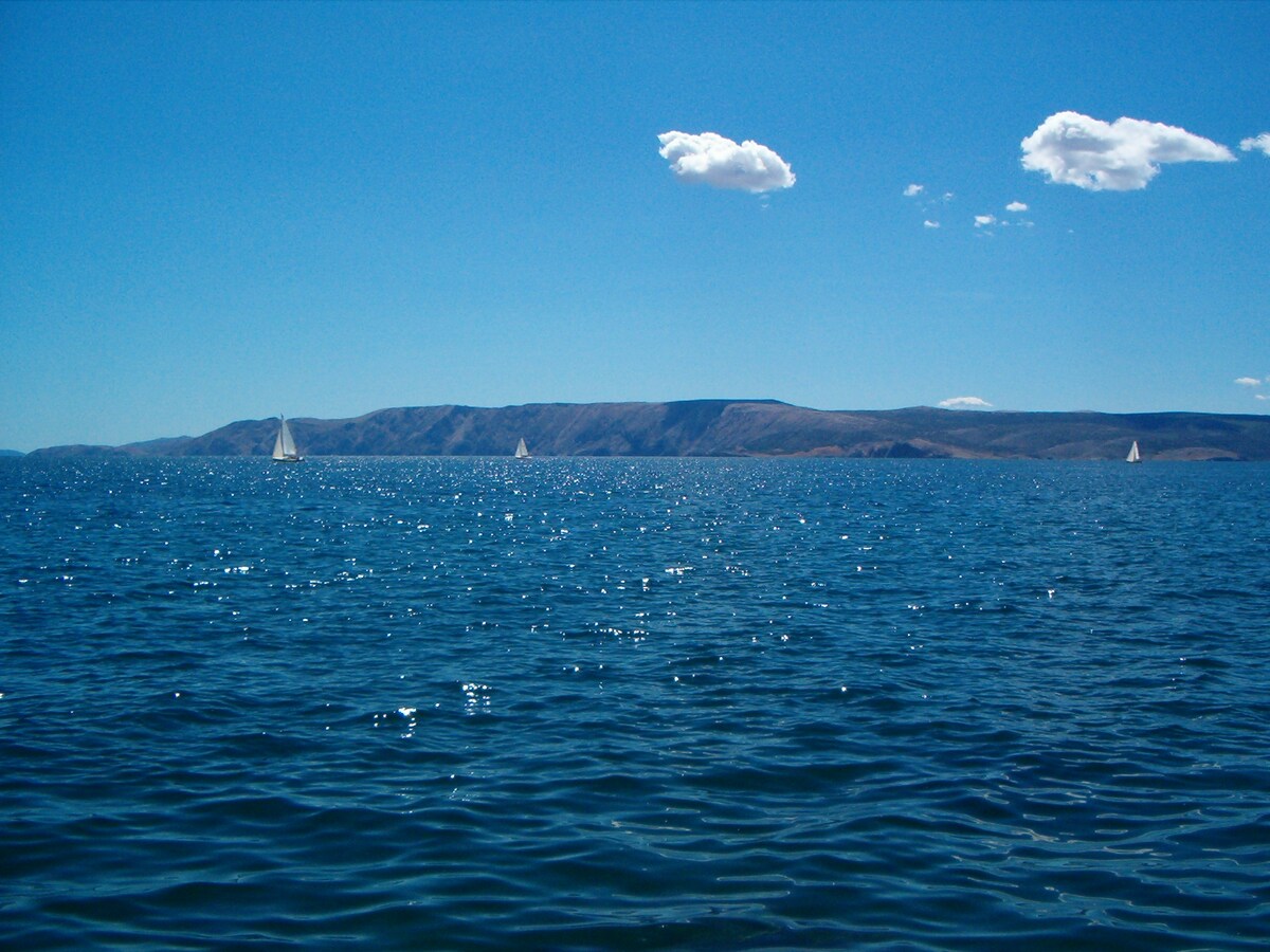 A serene view of the sea extends towards the horizon, displaying gentle waves reflecting sunlight. Sailboats are visible in the distance, framed against a clear blue sky with a few fluffy clouds. The backdrop consists of mountainous terrain, enhancing the peaceful coastal atmosphere.
