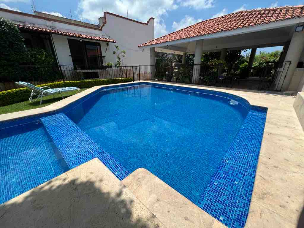 A private pool is featured, surrounded by natural stone and vibrant blue tiles. The pool area is shaded by a spacious palapa, and lush greenery borders the space. Clear skies and scattered clouds provide a bright atmosphere for relaxation.