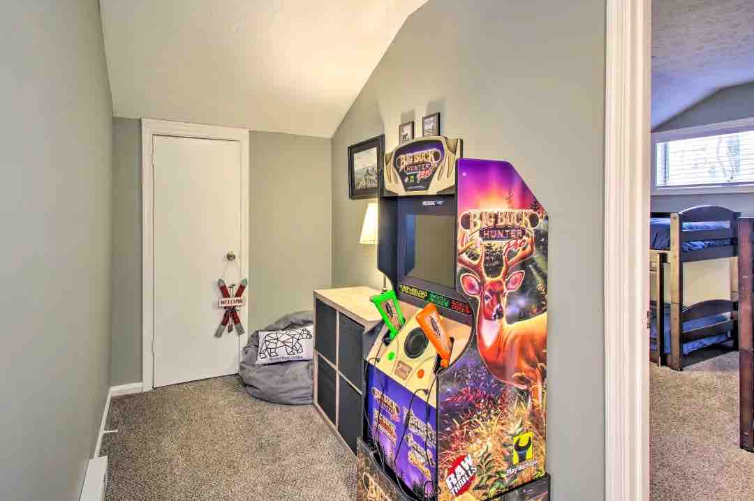 A cozy corner of the bunk room displays a colorful arcade game against a neutral wall. A soft bean bag is positioned beside a low storage unit. Framed photographs hang above the entertainment unit, and a door leads to another room.