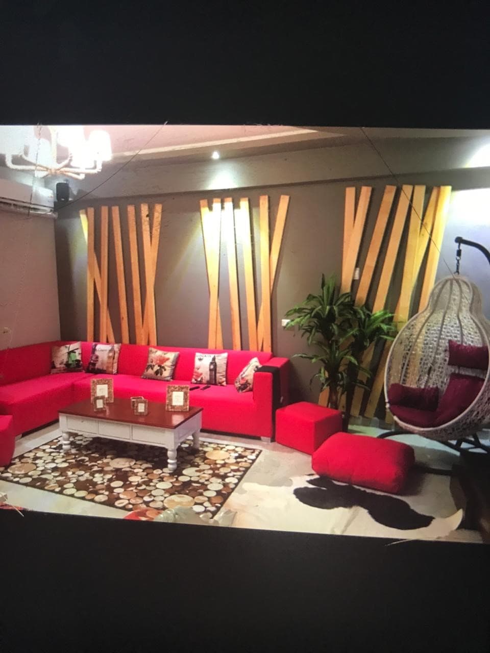 A lively living area is presented with a red sectional sofa complemented by decorative pillows. A hanging chair adds a unique touch, while a coffee table is positioned on a patterned rug. Textured wooden accents and a potted plant enhance the modern decor.