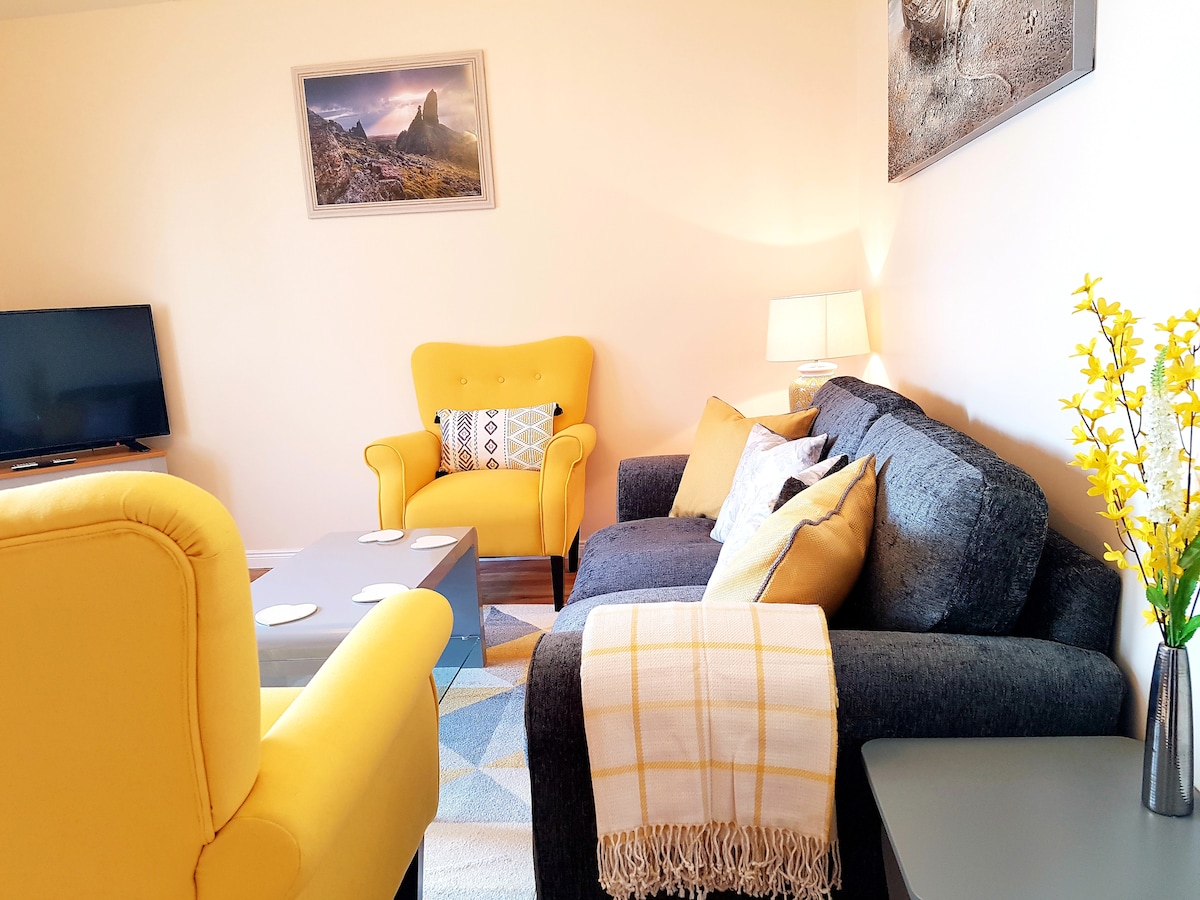 The lounge area features a gray sofa and two vibrant yellow armchairs, complemented by a wooden coffee table. A Smart TV is positioned across from the seating arrangement, while natural light enhances the space. Decorative flowers are arranged on a side table, adding a touch of color.