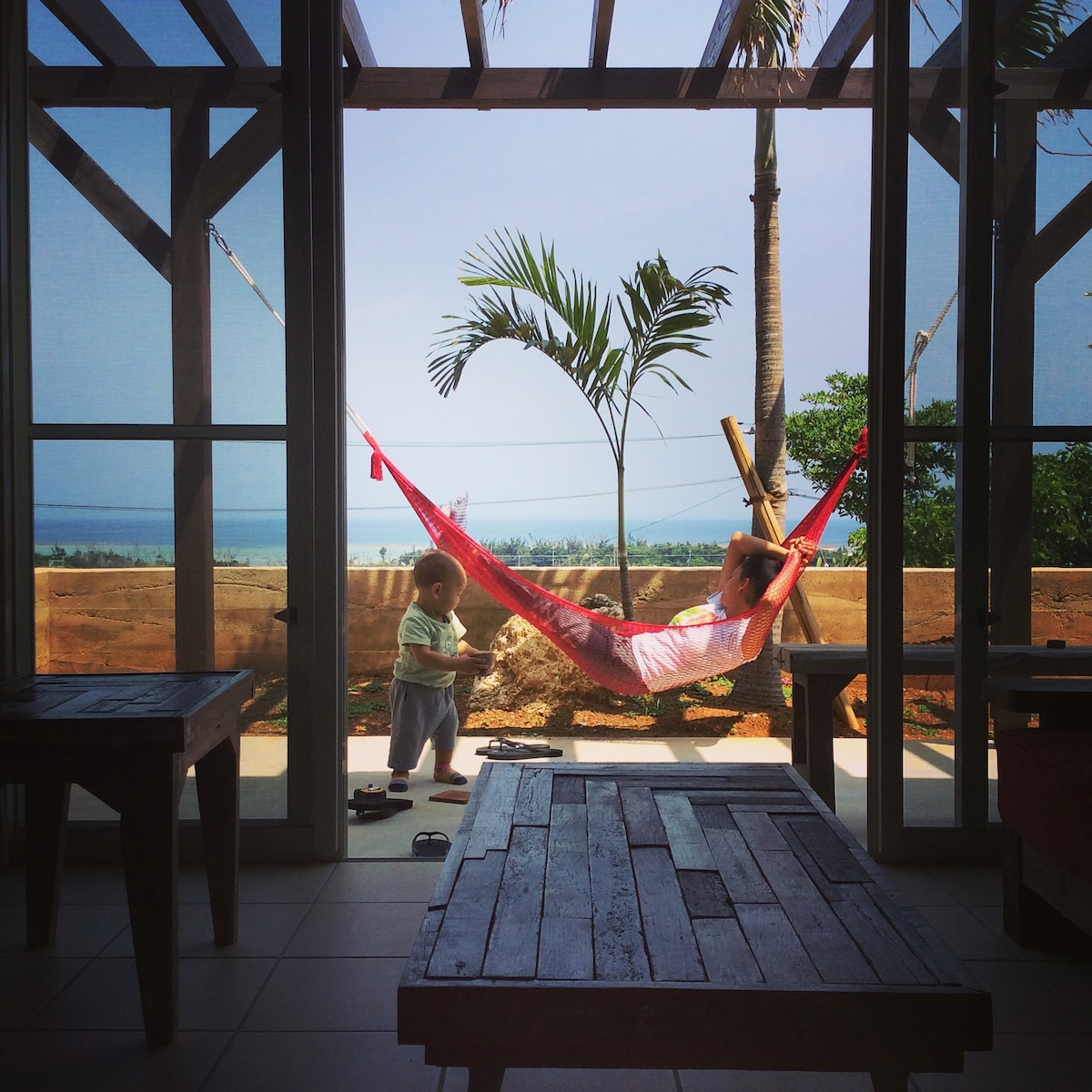 A hammock is stretched between pillars, offering a relaxing view of the ocean through large open windows. A child is seen playing nearby on the tiled floor, while green palm trees frame the scene. Natural light floods the space, enhancing the tranquil ambiance.
