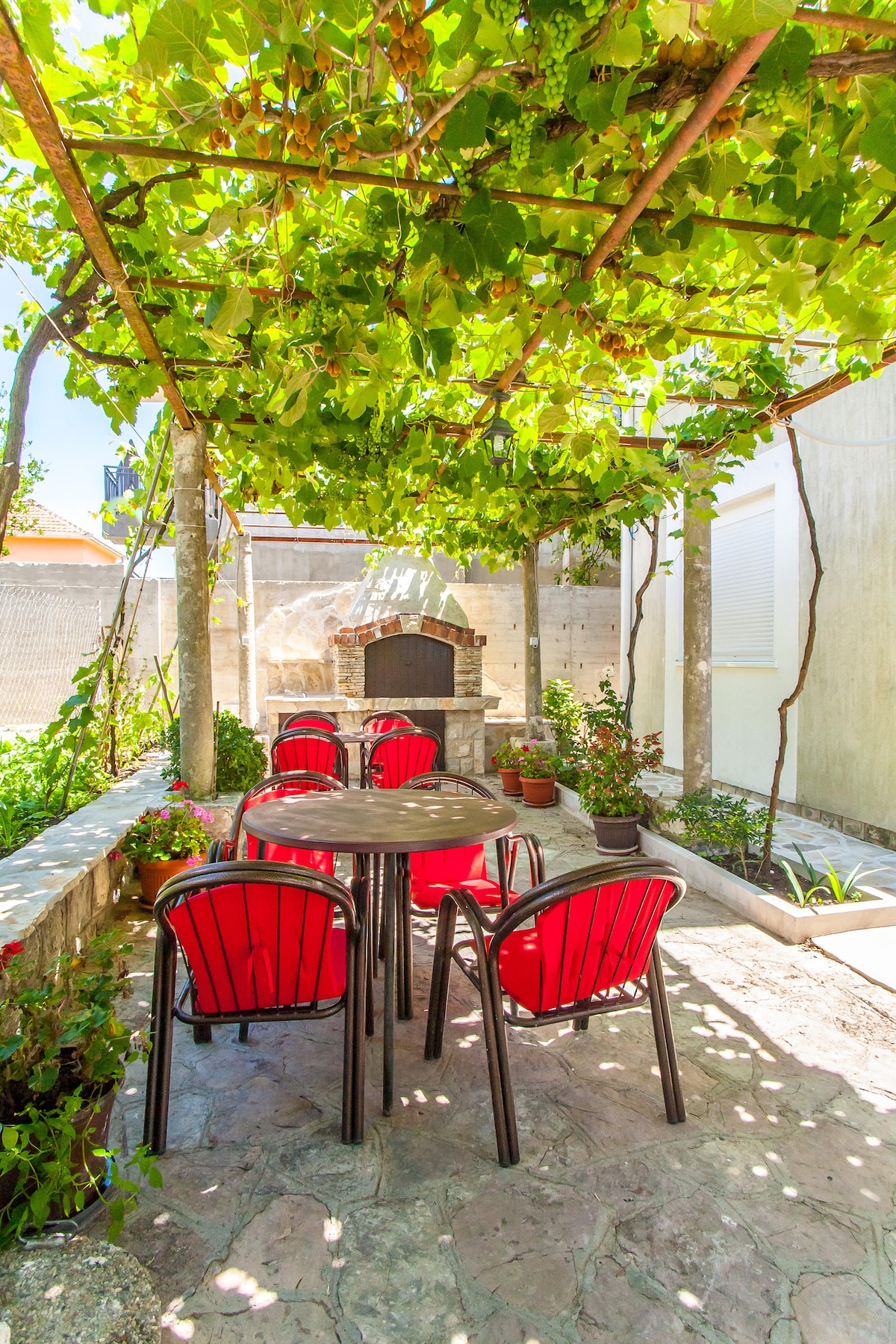 A shaded outdoor seating area is presented under a canopy of green vines. A round table is surrounded by six red chairs, with planters featuring various plants positioned around the space. A barbecue grill is visible in the background, enhancing the area’s functionality.