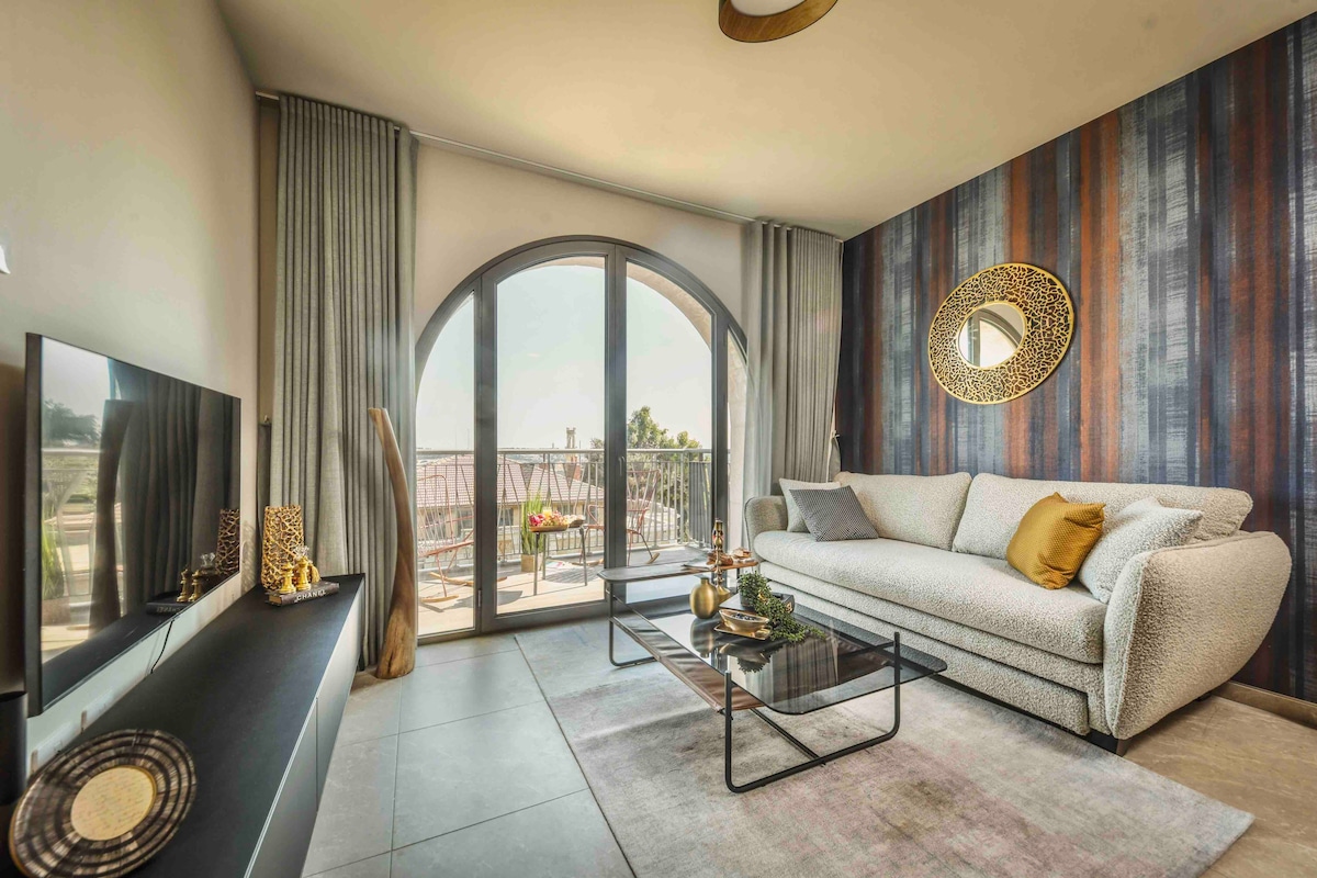 A bright living room features a large, curved glass door leading to a balcony. A plush seating area is arranged with a light-colored sofa adorned with decorative pillows. A low coffee table sits on a soft area rug, while a stylish mirror complements the contemporary decor.