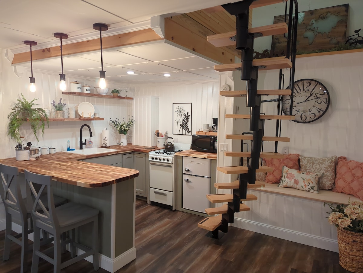 The compact kitchen area features modern appliances, a wooden countertop, and stool seating. A spiral staircase leads to an upper level. Natural light illuminates the space, highlighting decorative plants and wall art, creating a welcoming ambiance.