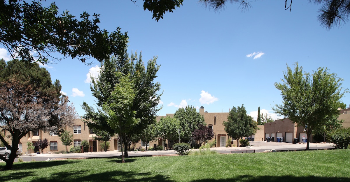 A scenic view of a well-maintained neighborhood is presented, featuring a grassy area with trees on the forefront. In the background, two-story condo units are visible, showcasing their natural landscaping and the inviting blue sky above.
