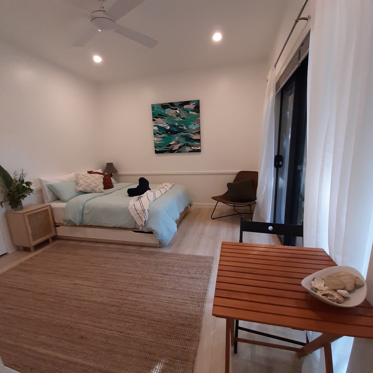 A modern studio is presented, featuring a spacious bed adorned with a light blue blanket and several decorative pillows. A small wooden table with a bowl of shells is positioned nearby, alongside a seating area. Natural light floods the space through glass doors, complemented by light curtains.