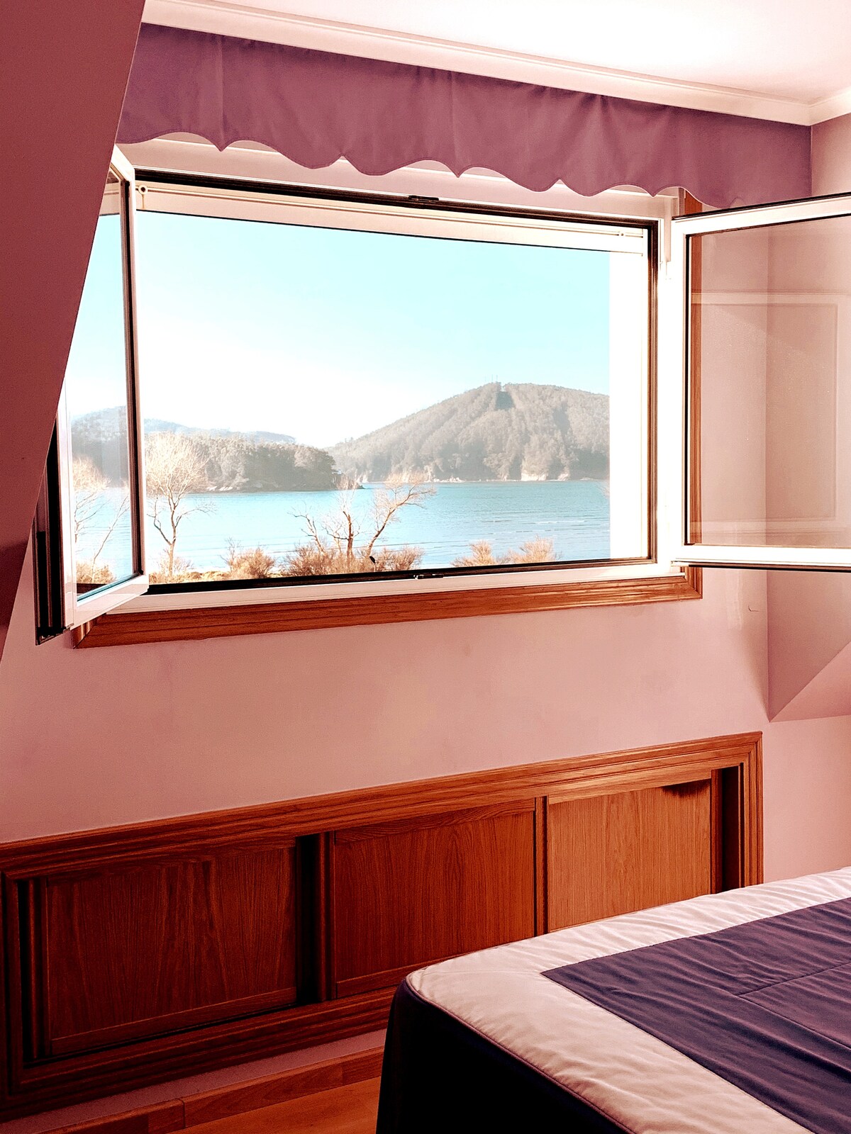 A bright bedroom features a large open window, allowing natural light to illuminate the space. A view of rolling hills and water is visible outside, framed by wooden elements. The bed is positioned near the window, enhancing the serene atmosphere.