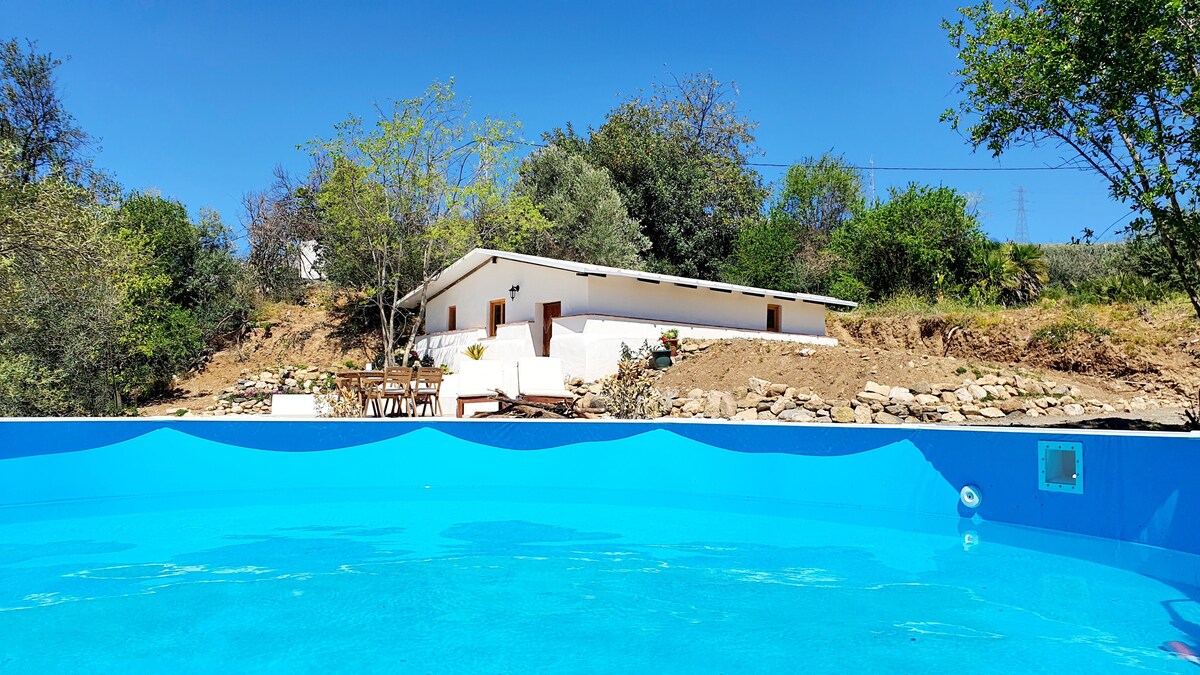 A vibrant blue swimming pool is showcased in the foreground, with a spacious white accommodation structure partially visible in the background. The surrounding landscape features greenery and natural vegetation under a clear blue sky.