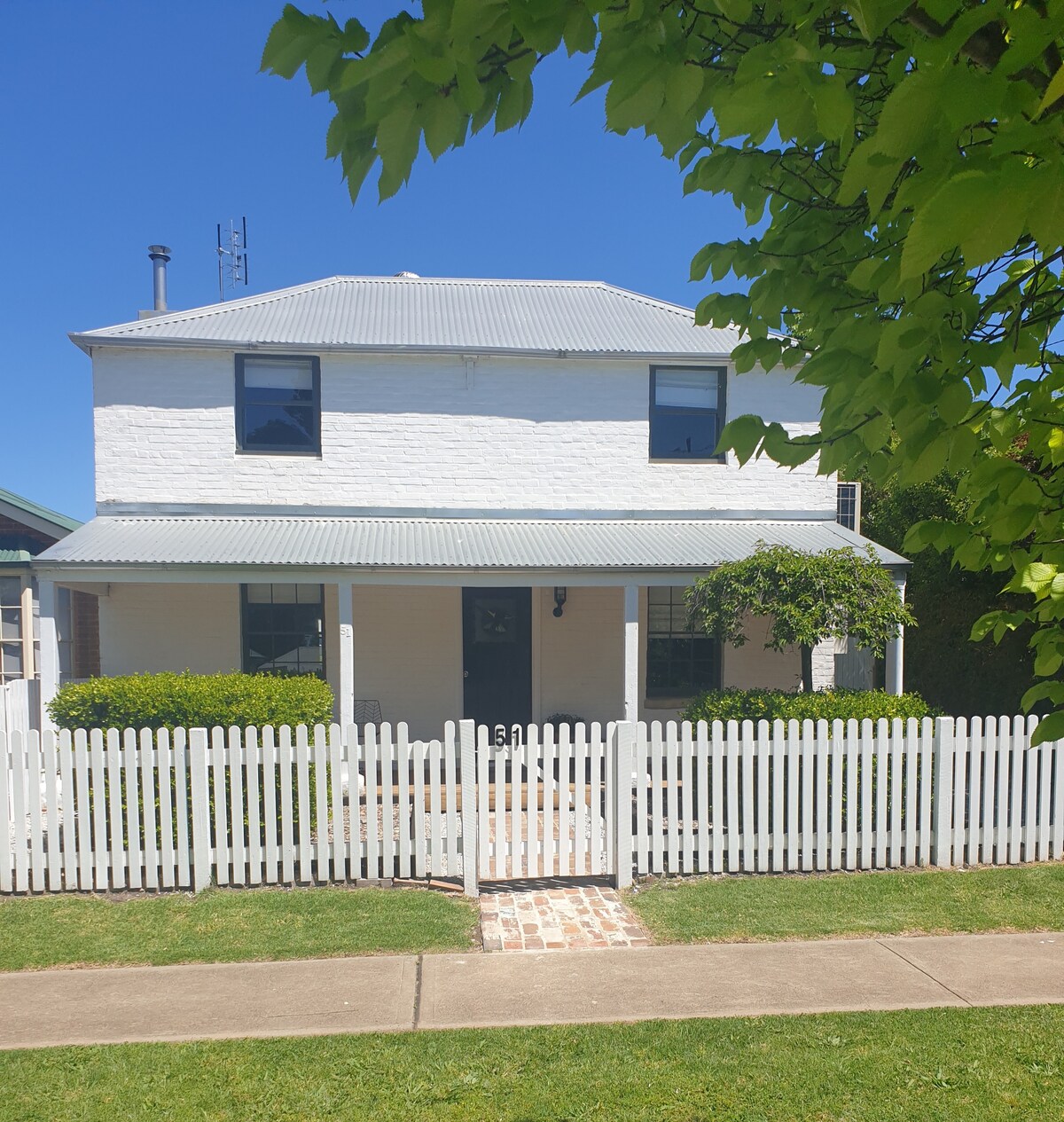 The two-story cottage features a classic design with white brick exterior and a metal roof. A charming white picket fence encloses the front yard, complemented by a pathway of bricks leading to the entrance. Lush greenery provides a welcoming touch.